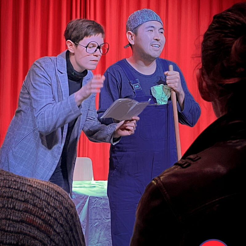 eine Frau liest auf einer Bühne aus einem Heft. Sie steht neben einem Mann, welcher im blauen Overall einen Besen hält.