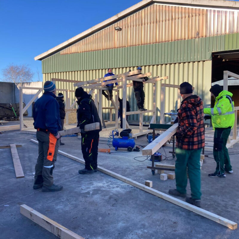 Sieben Männer bauen gemeinsam eine Bude aus Holzlatten vor einem Lagergebäude im Winter.