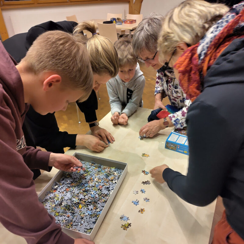 zwei Kinder und drei Frauen puzzeln gemeinsam um einen Tisch herum.