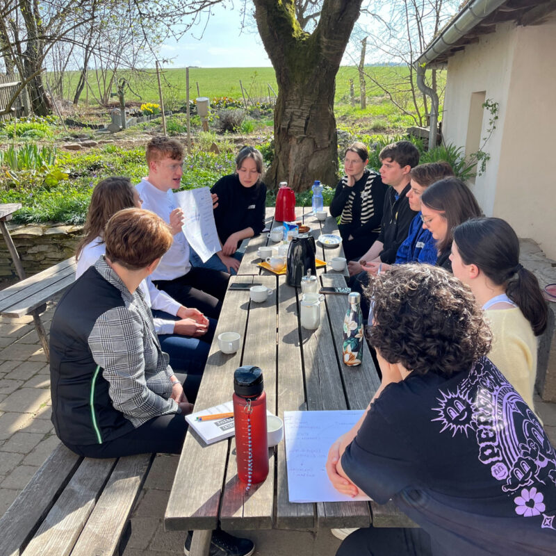 Junge Menschen sitzen um einen Tisch draußen und diskutieren gemeinsam.