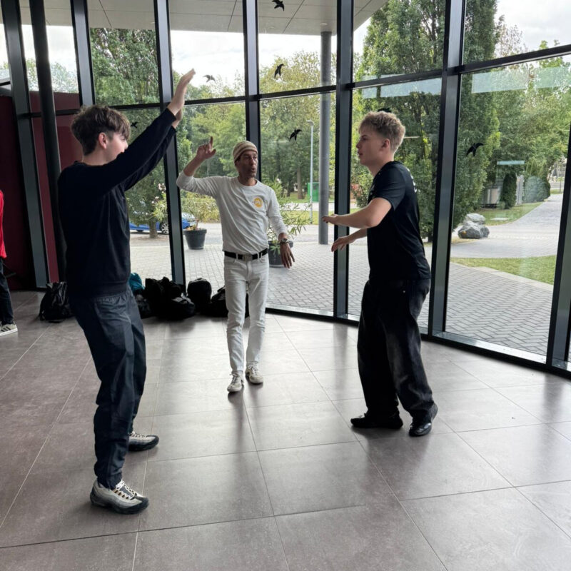 „Drei Jugendliche üben mit einem Kursleiter Bewegungen in einem hellen Raum mit großen Fenstern