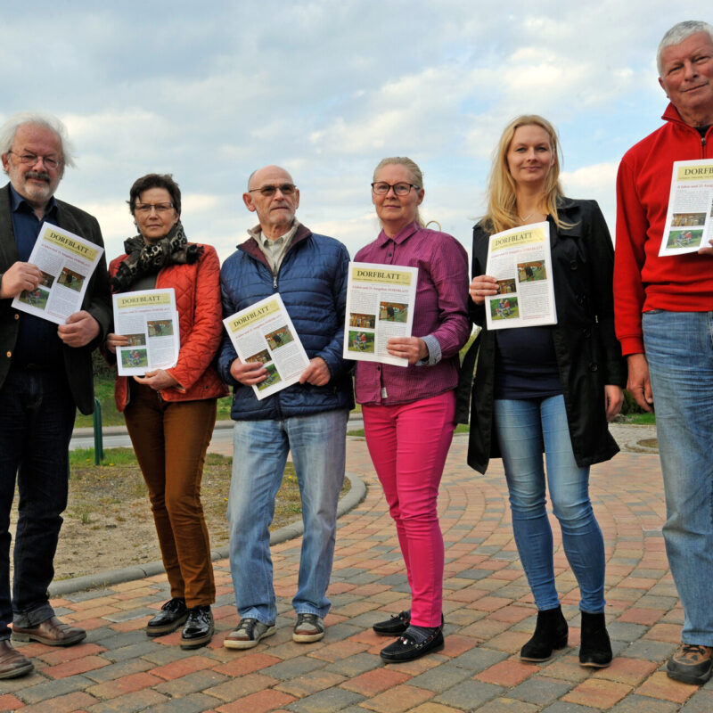 Sechs Erwachsene stehen nebeneinander im Freien auf einem gepflasterten Platz. Jede Person hält ein Exemplar des „Dorfblatt“-Newsletters in der Hand. Die Gruppe lächelt in die Kamera; im Hintergrund ist der Himmel leicht bewölkt