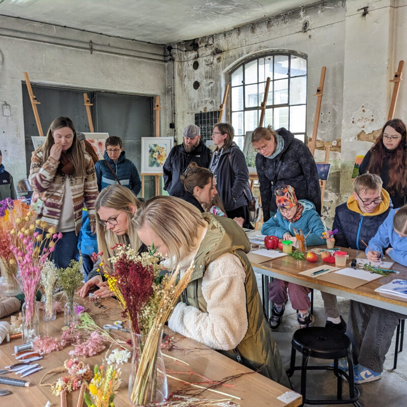 „Gruppe von Menschen arbeitet an einem Werk- und Kreativtisch mit Trockenblumen und Materialien; im Hintergrund stehen weitere Teilnehmende und betrachten Bilder an Staffeleien.“