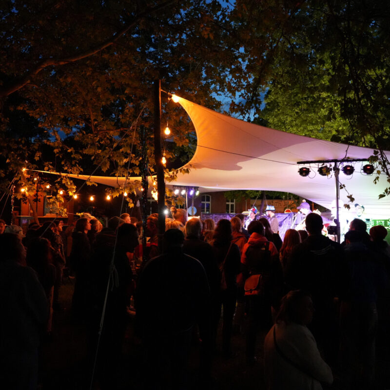 Menschen stehen abends unter beleuchteten Bäumen vor einer kleinen Bühne mit Live-Musik bei einer Open-Air-Veranstaltung.