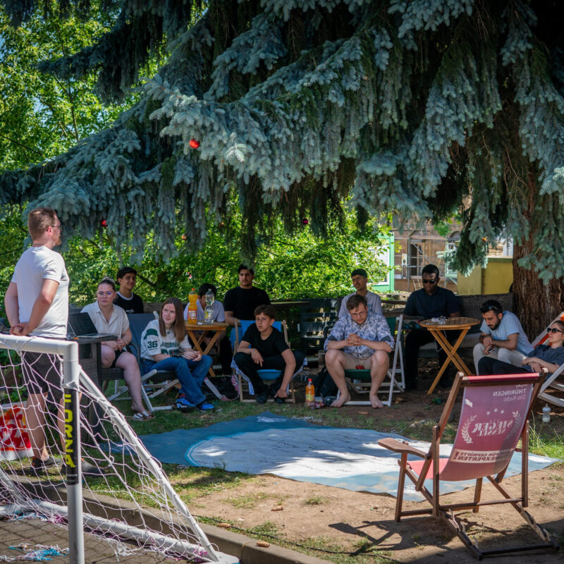 „Gruppe junger Menschen sitzt im Freien unter einem Baum im Stuhlkreis und nimmt an einem Workshop teil.“