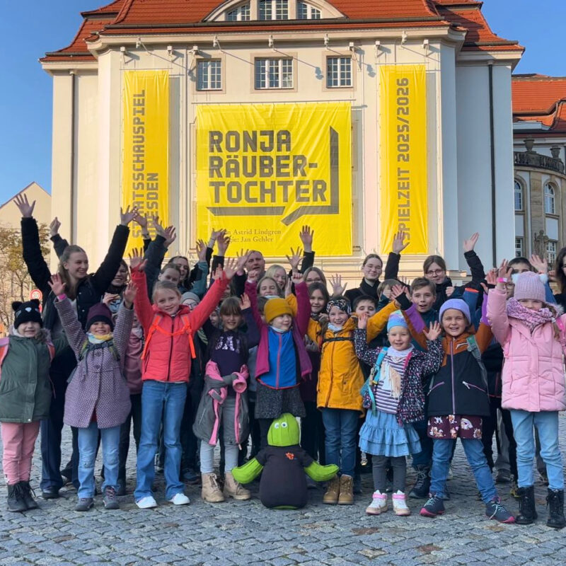Eine Gruppe Kinder und junger Erwachsener steht vor dem Schauspielhaus Dresden und winkt in die Kamera