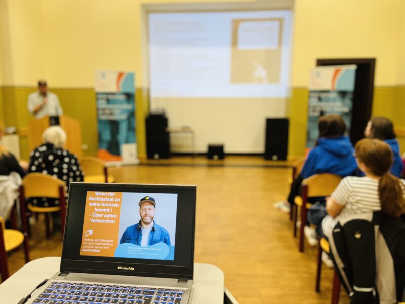 Gruppe in Schulaula hört einem Vortrag zu.