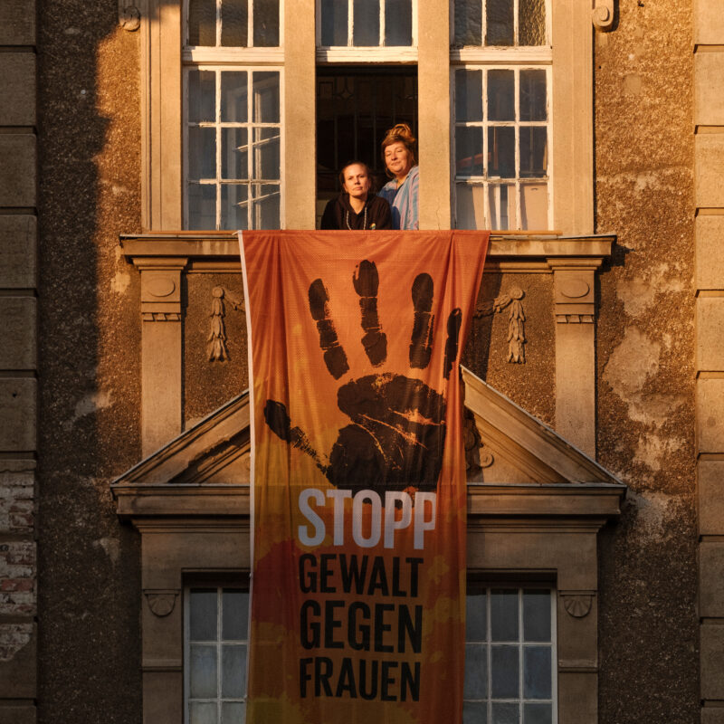 Zwei Frauen schauen bei Abendlicht aus einem Gebäudefenster, unter ihnen hängt an der Fassade ein großes Banner mit der Aufschrift "STOPP Gewalt gegen Frauen)"