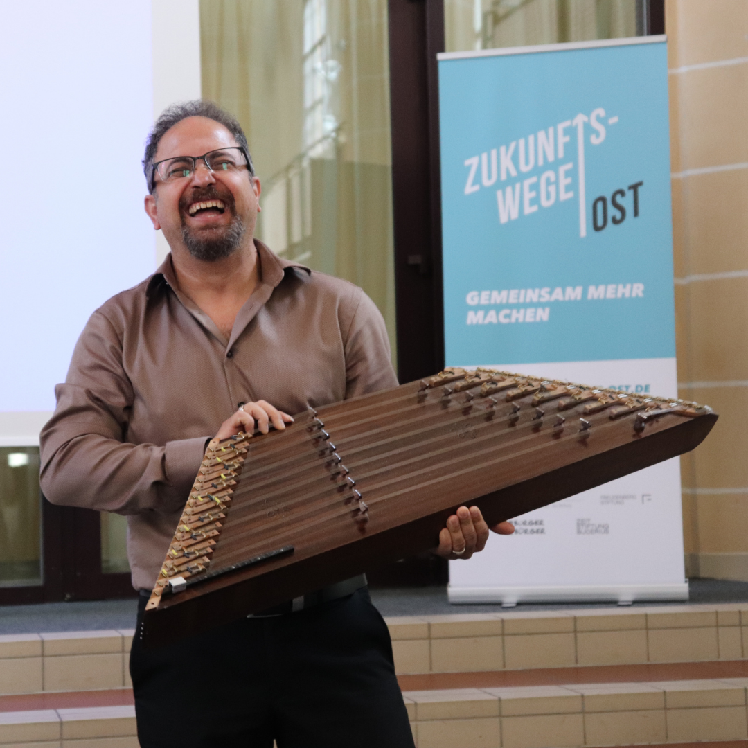 Ein Mann hält das Musikinstrument Shantu präesntierend hoch und lacht.