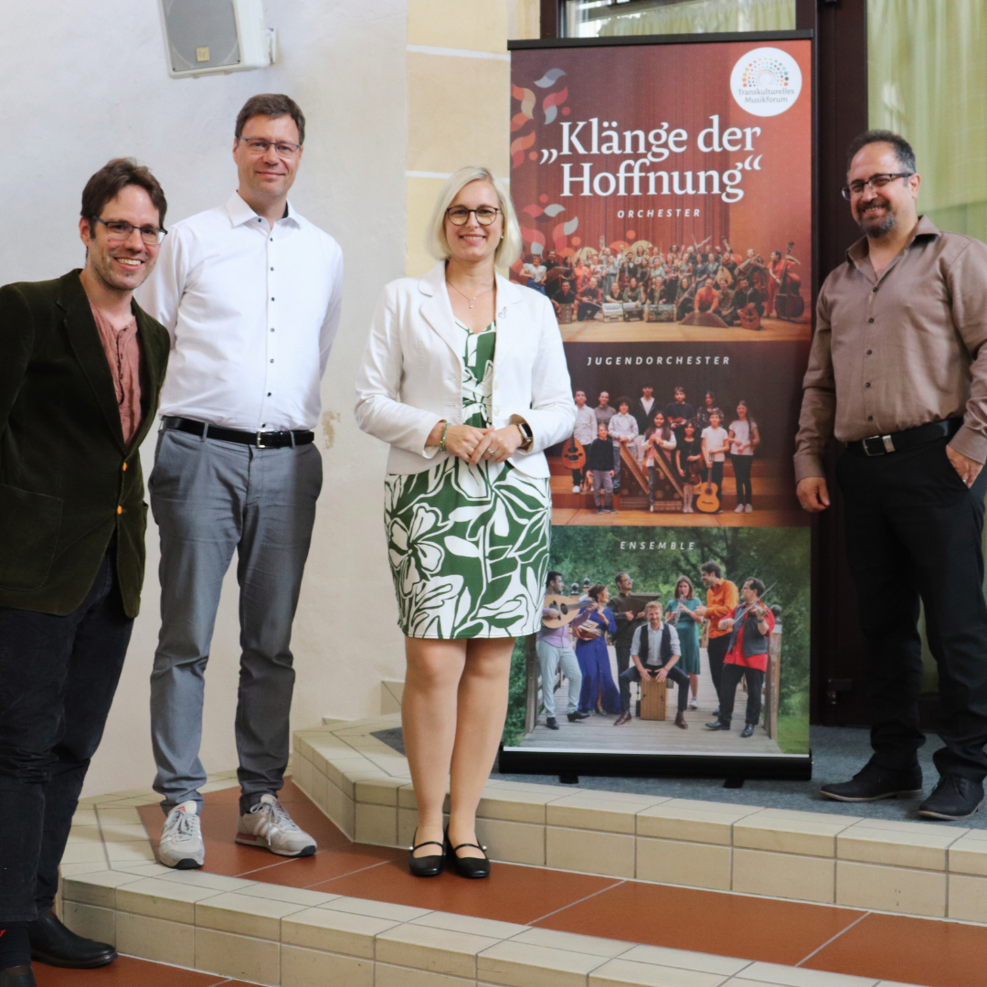 Vier Menschen stehen vor einem Aufsteller mit der Aufschrift: "Klänge der Hoffnunf" Orchester und schauen freundlich.