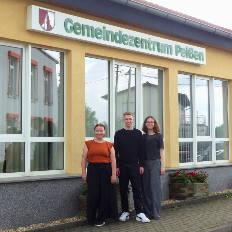 Drei junge Menschen stehen draußen vor einem Gebäude auf dem steht "Gemeindezentrum Peißen"
