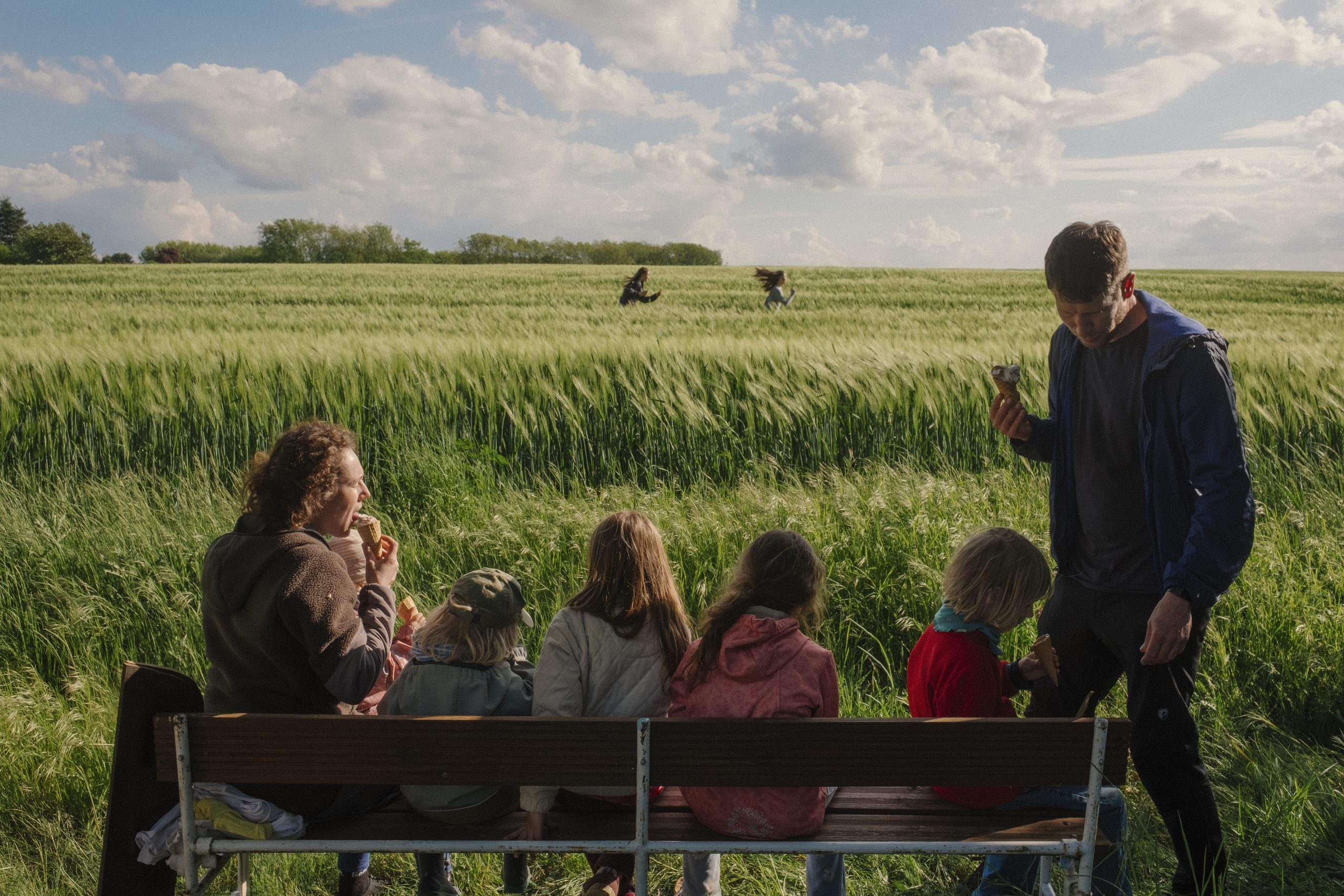 Eltern sitzen mit ihren Kindern auf einer Bank vor einem grünen Feld. Es sieht sehr ländlich aus, der Himmel ist blau.