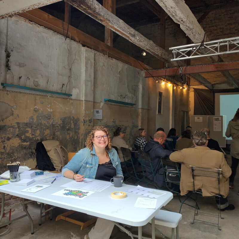 Person sitzt an einem Anmeldetisch in einer Industriehalle, im Hintergrund nehmen Teilnehmende auf Stühlen Platz.
